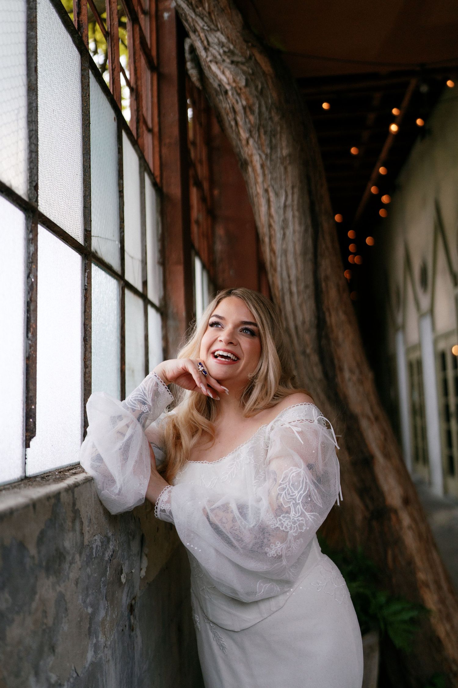 Radiant blonde bride in off-shoulder lace wedding dress laughing joyfully beside large windows with string lights.