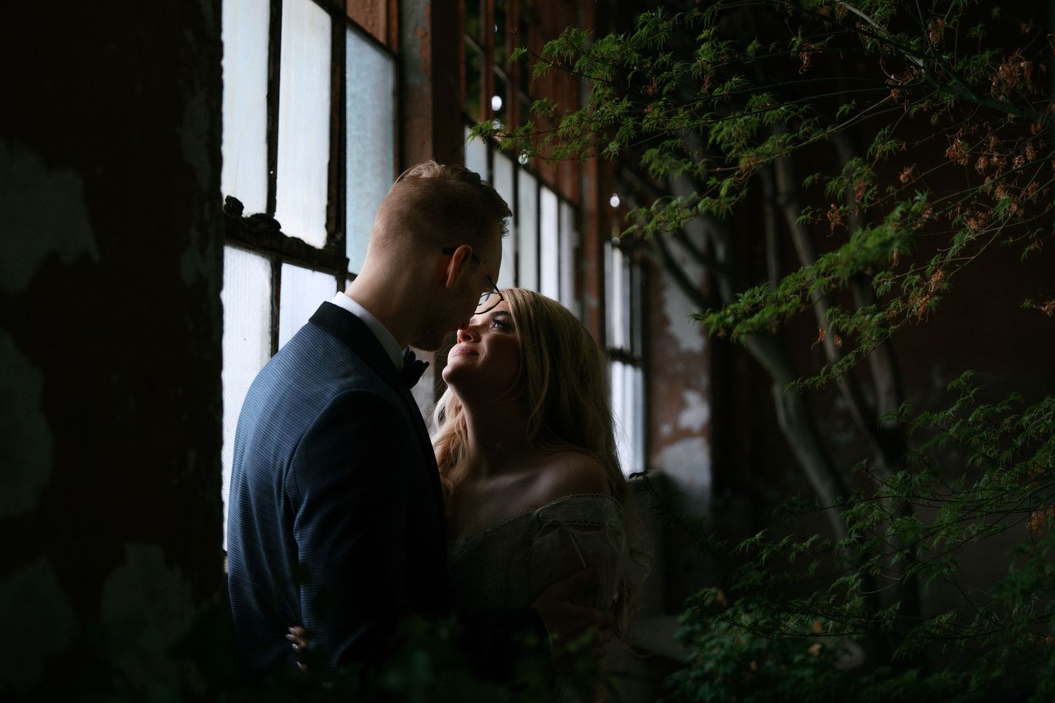 Groom tenderly kisses bride near large industrial window surrounded by lush green plants in a moody, romantic setting.