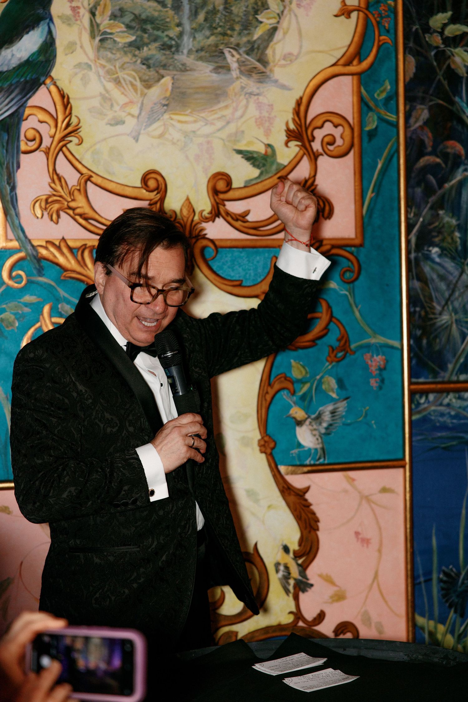 Man in tuxedo holding microphone with arm raised at elegant wedding reception.