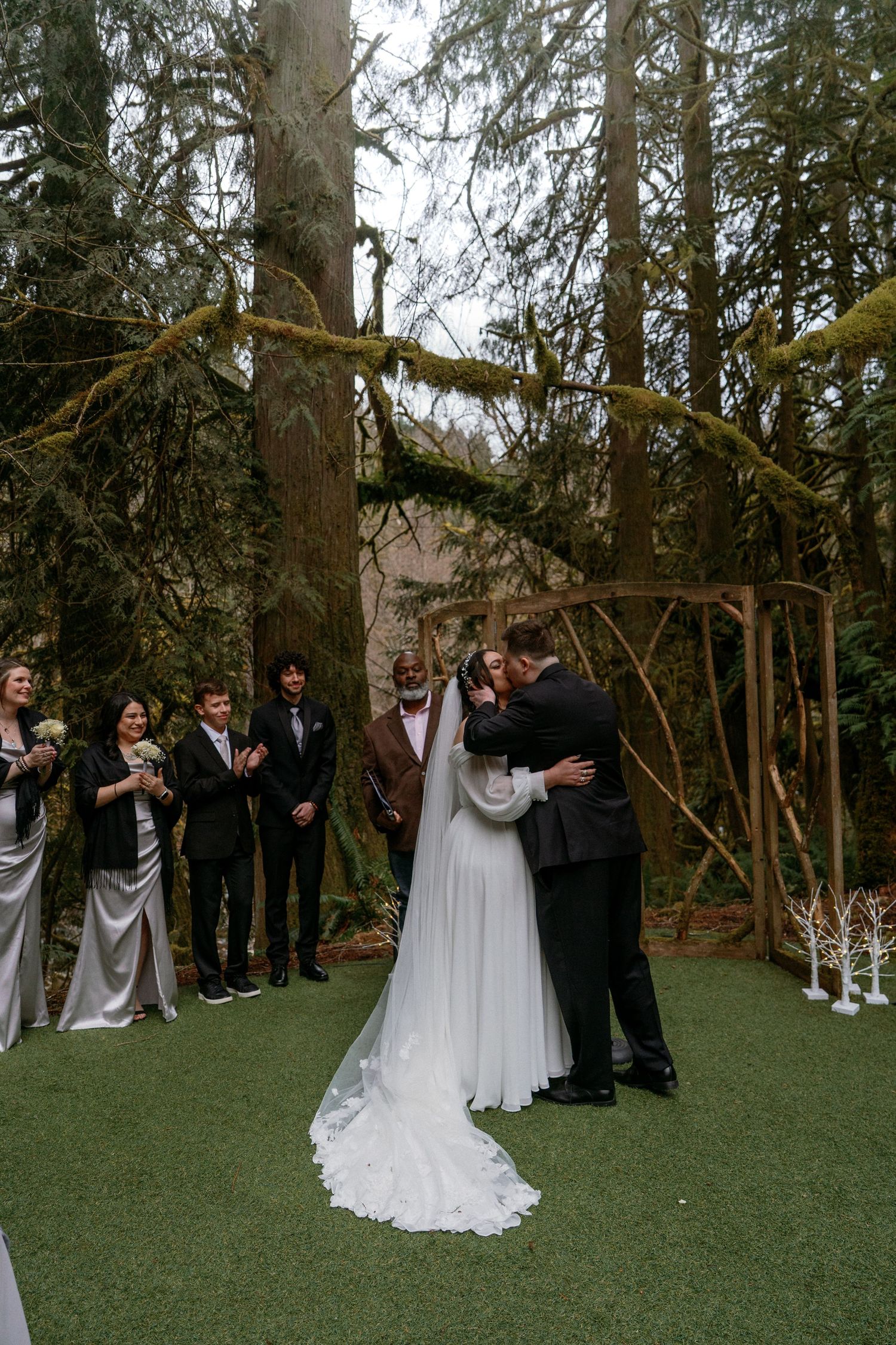 Bride and groom share first kiss under wooden arch at emotional outdoor forest wedding ceremony.