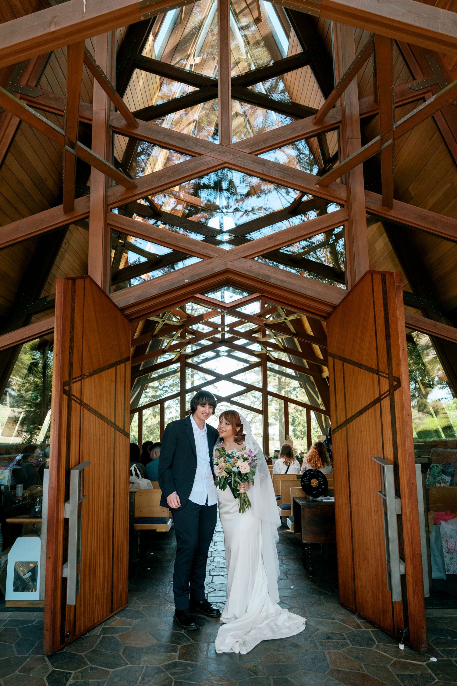 Bride and groom standing together under geometric wood arch in a glass chapel.