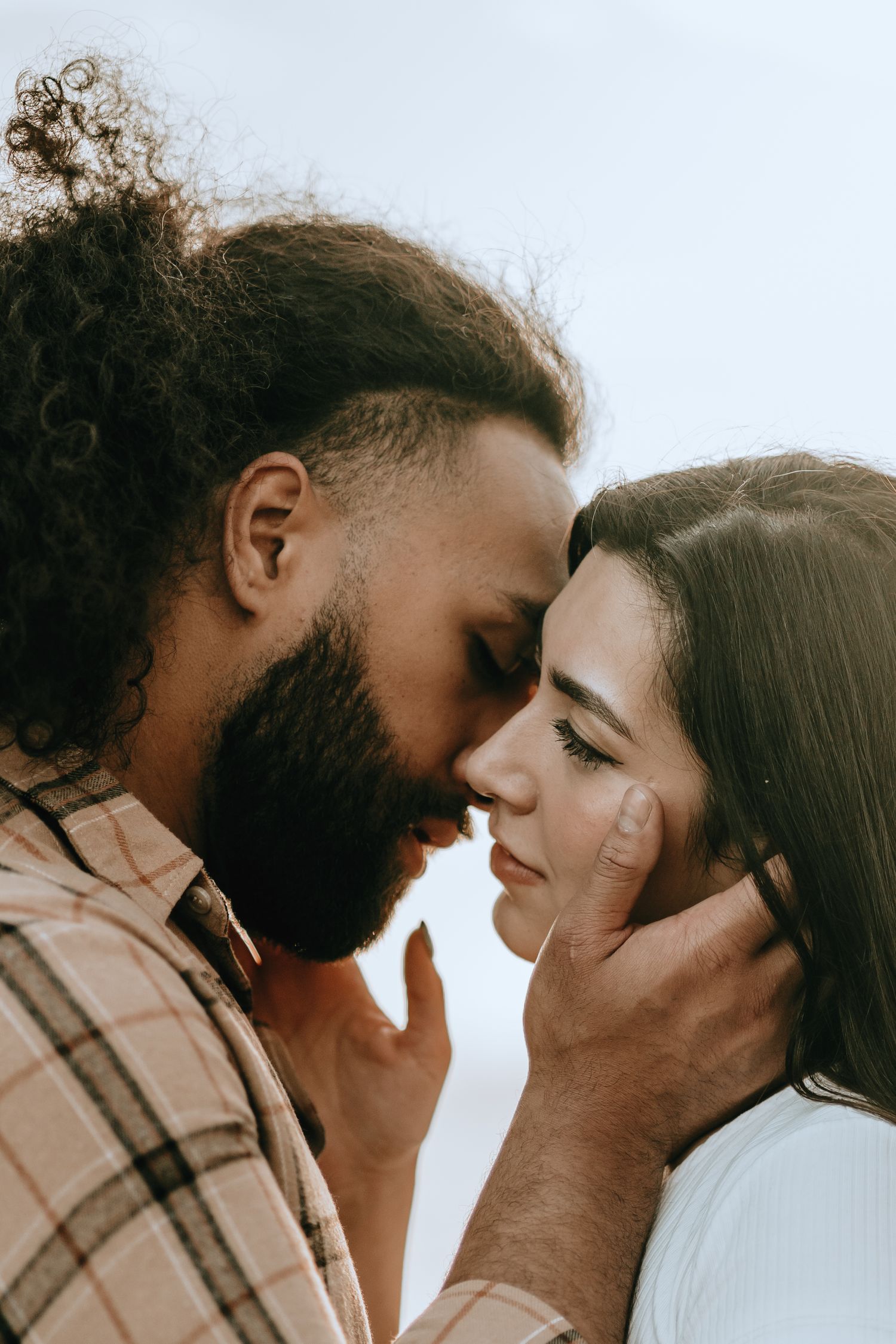Romantic couple with foreheads touching in an intimate close-up moment against a soft sky background.