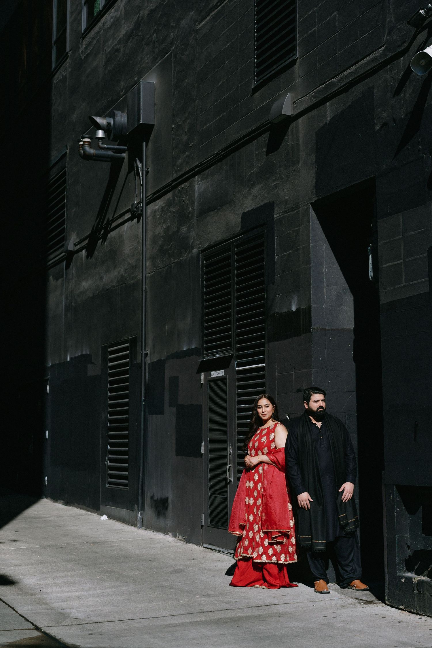 Couple in traditional South Asian attire posing against a dark industrial building wall in an urban setting.