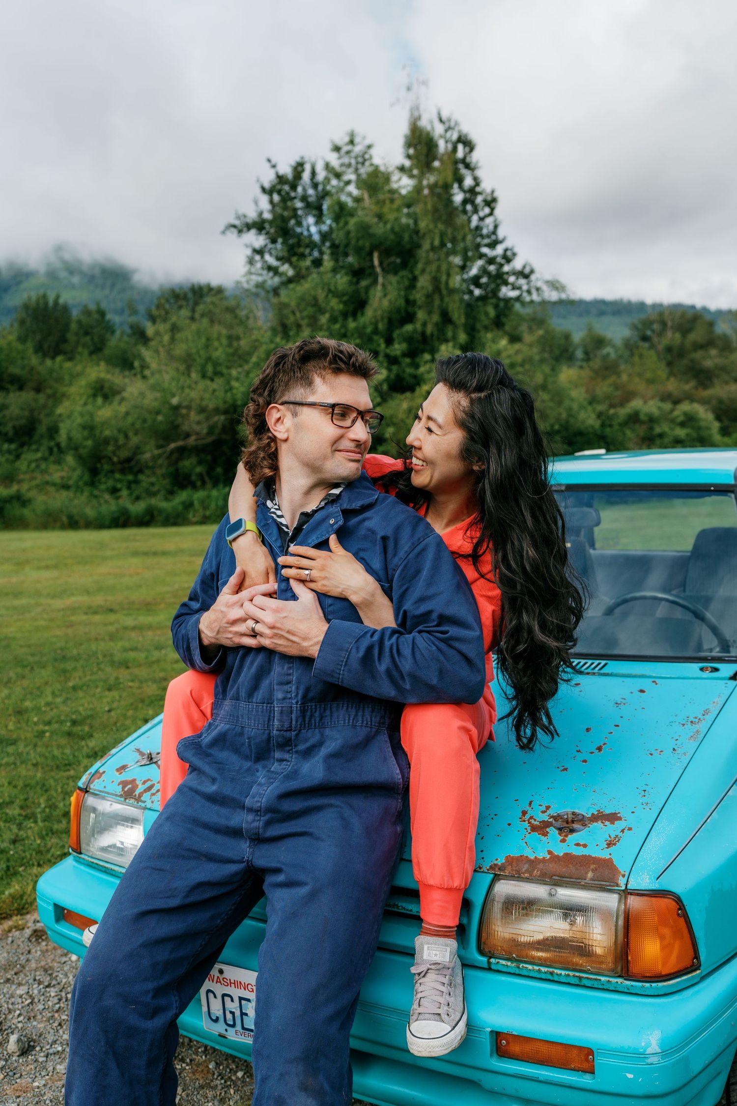 A couple embraces affectionately on the hood of a vintage turquoise car in a green landscape.