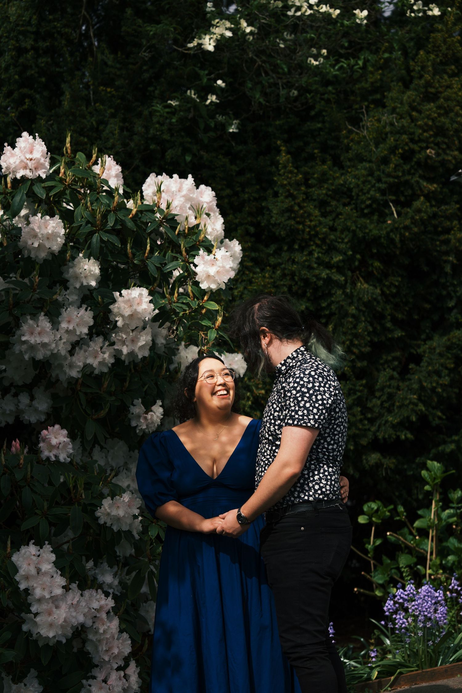 Joyful couple holding hands and laughing beside white rhododendron blooms in a garden setting.