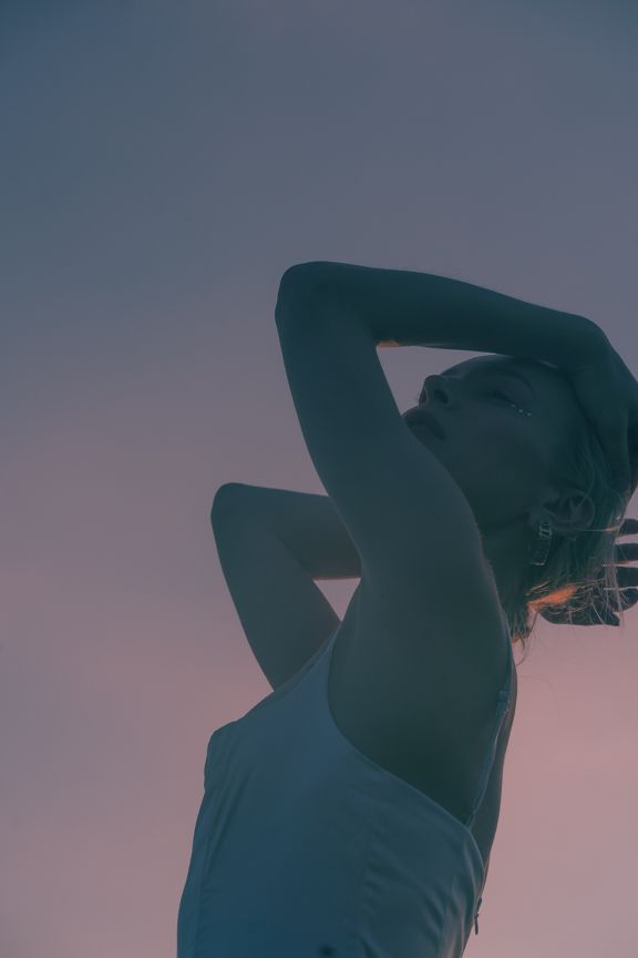 Silhouette of woman in white dress with arms raised against pink and purple sunset sky.