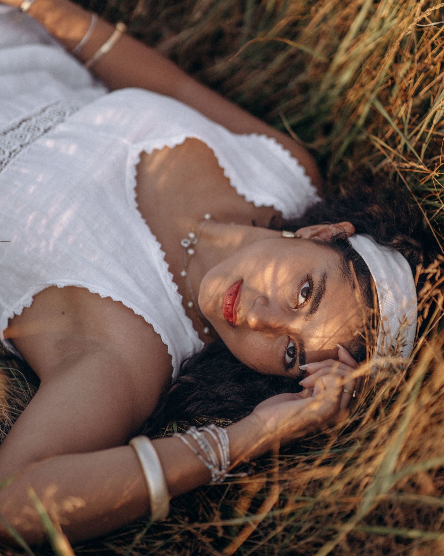 Woman in white dress lying in golden grass, wearing headband and bracelets, gazing at camera.