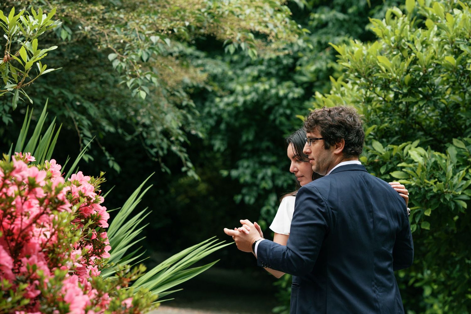 Two men in navy suits standing near pink flowers and tropical plants in a lush garden setting.