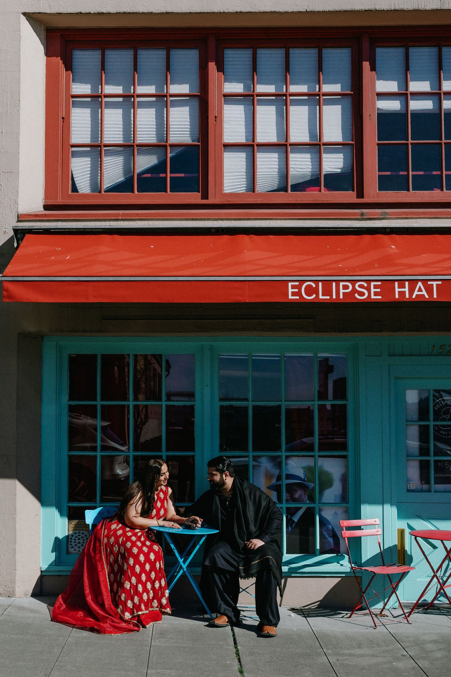 Bride in red Indian bridal dress and groom in black sit outside Eclipse Hat shop in bright sunlight.