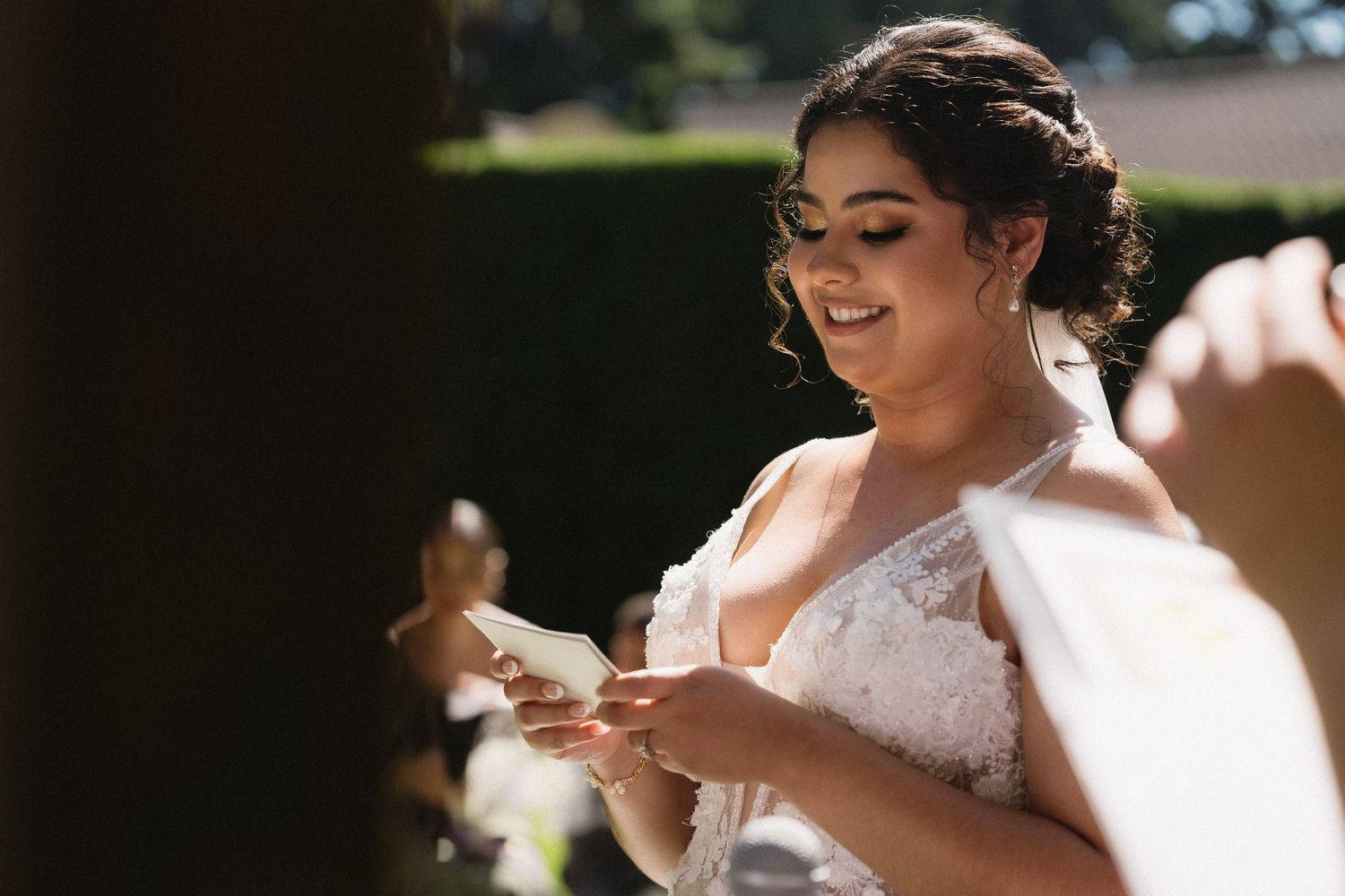 Radiant bride in lace wedding gown smiles while reading vows card in warm sunlight outdoors.