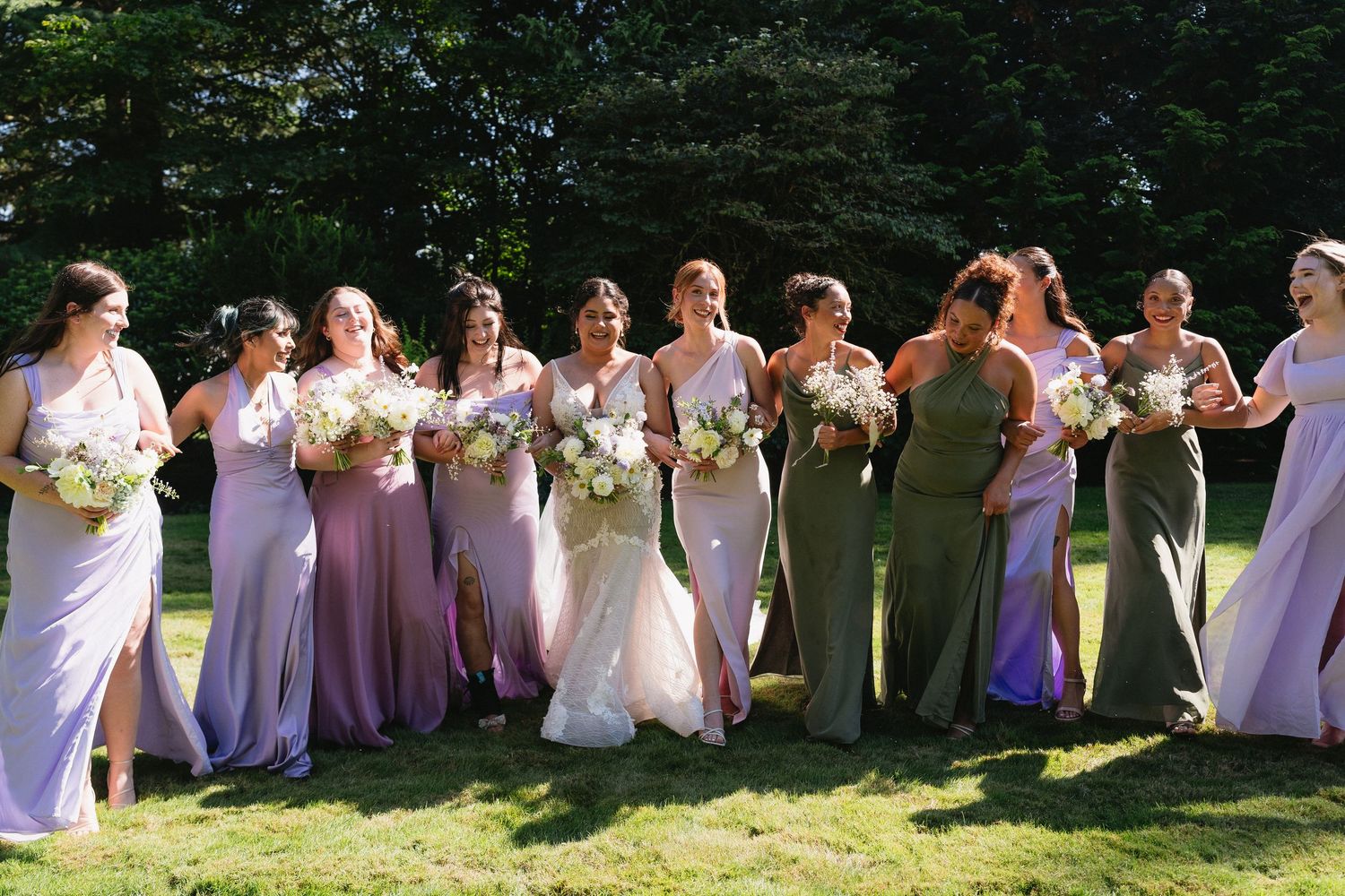 Bride in lace gown poses with bridesmaids in mixed purple and sage green dresses outdoors.