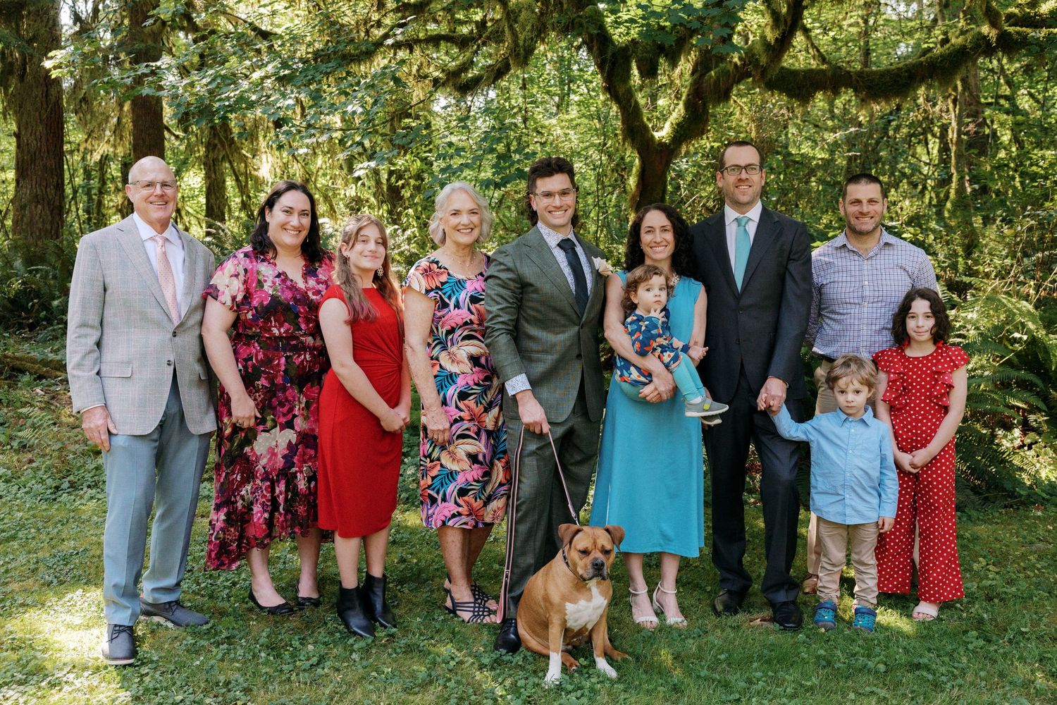 Wedding family group portrait with dog on a sunny green lawn surrounded by trees.