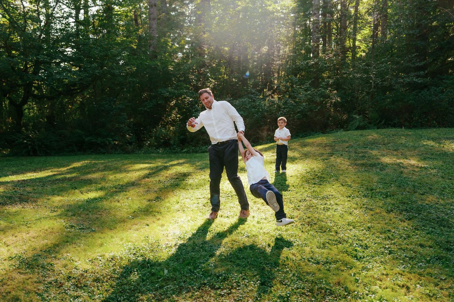 Man in white shirt joyfully swings a child by the arms on a sunny green lawn at outdoor wedding.