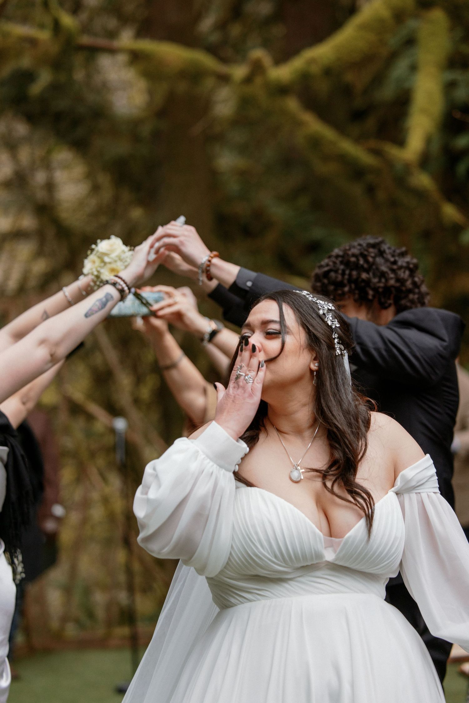 Radiant bride in white gown blowing a kiss during celebratory outdoor forest wedding recessional.