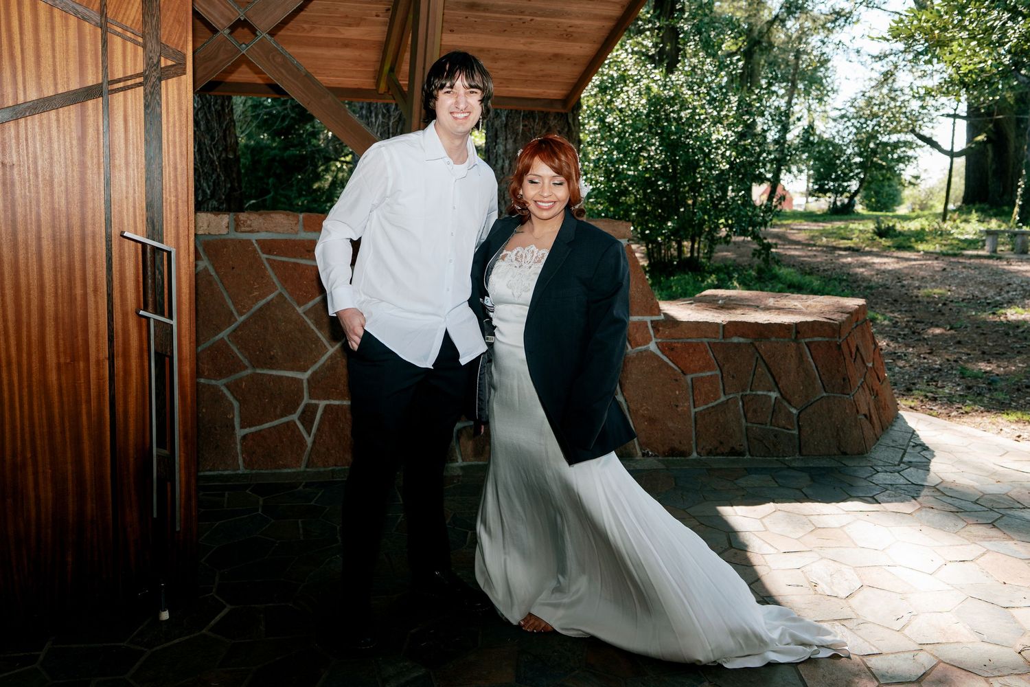 Bride and groom posing outdoors near stone wall in casual post-wedding portrait.