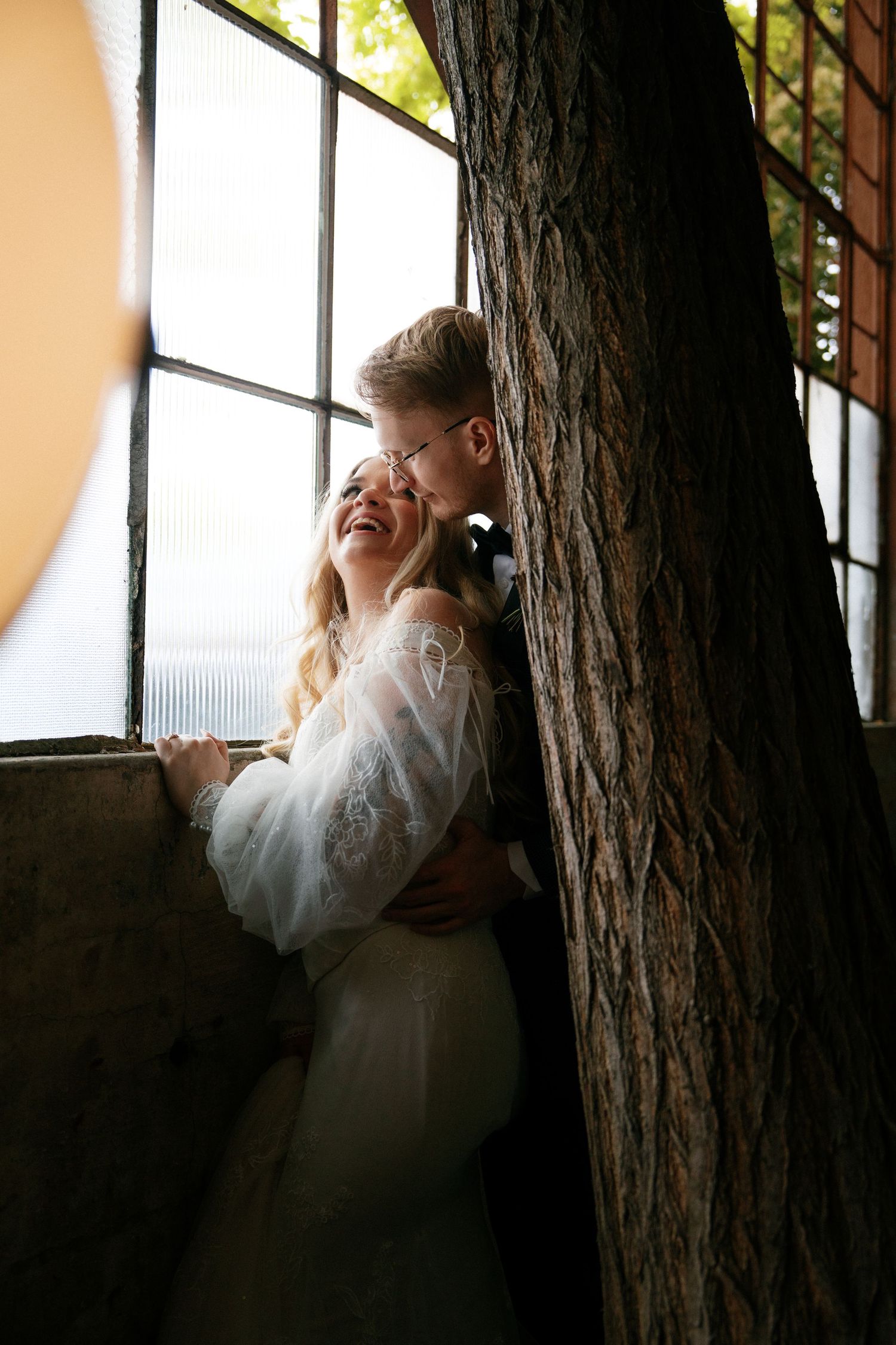 Laughing bride in lace gown embraced by groom beside a large tree trunk near industrial windows in rustic venue.