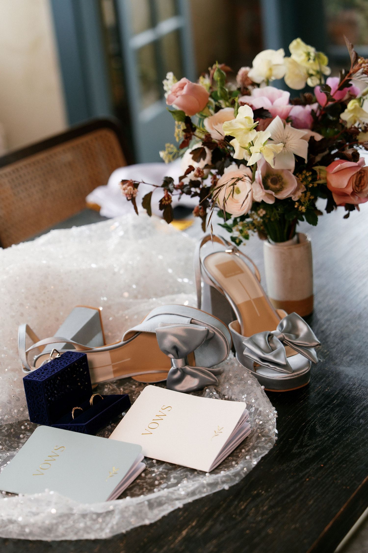 Wedding flat lay featuring silver heels, vow books, rings, and floral bouquet on table.