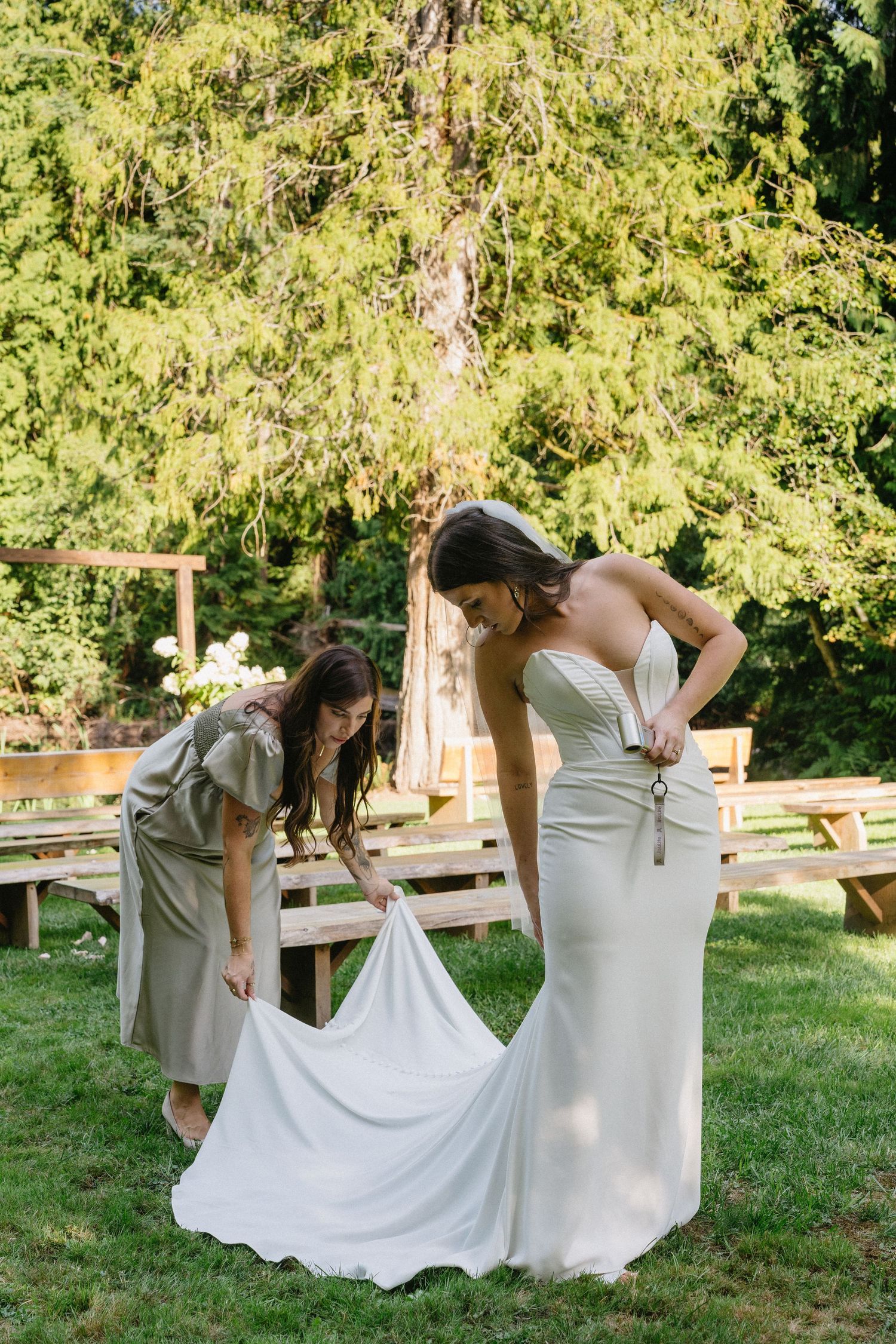 Bridesmaid helping bride adjust her white wedding gown outdoors at a garden ceremony venue.