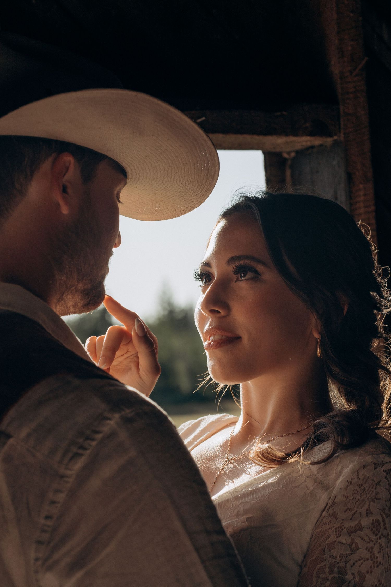Bride gazes up lovingly at groom wearing white cowboy hat in soft golden backlit barn setting.