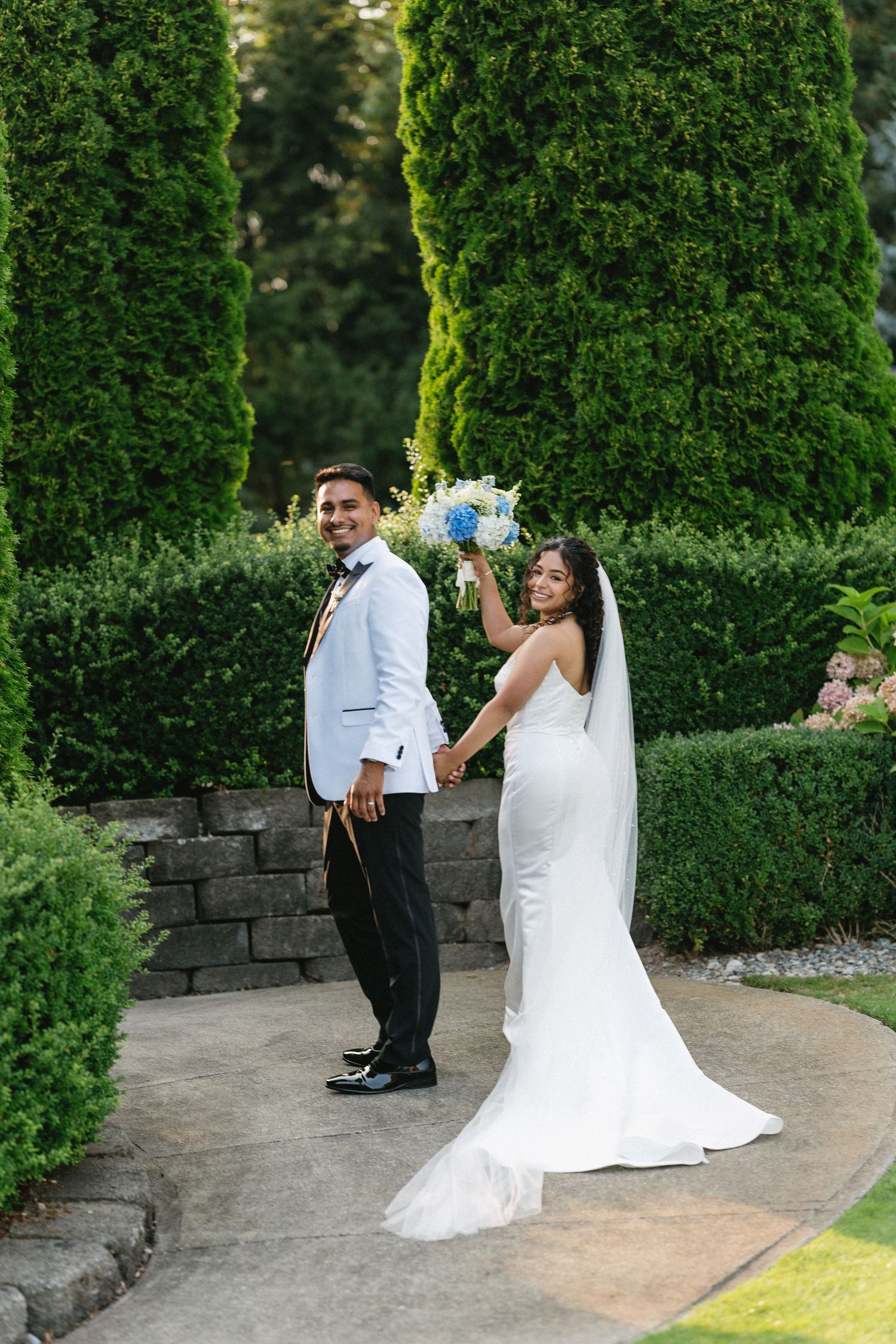 Bride and groom pose in lush garden holding bouquet among tall cypress trees.
