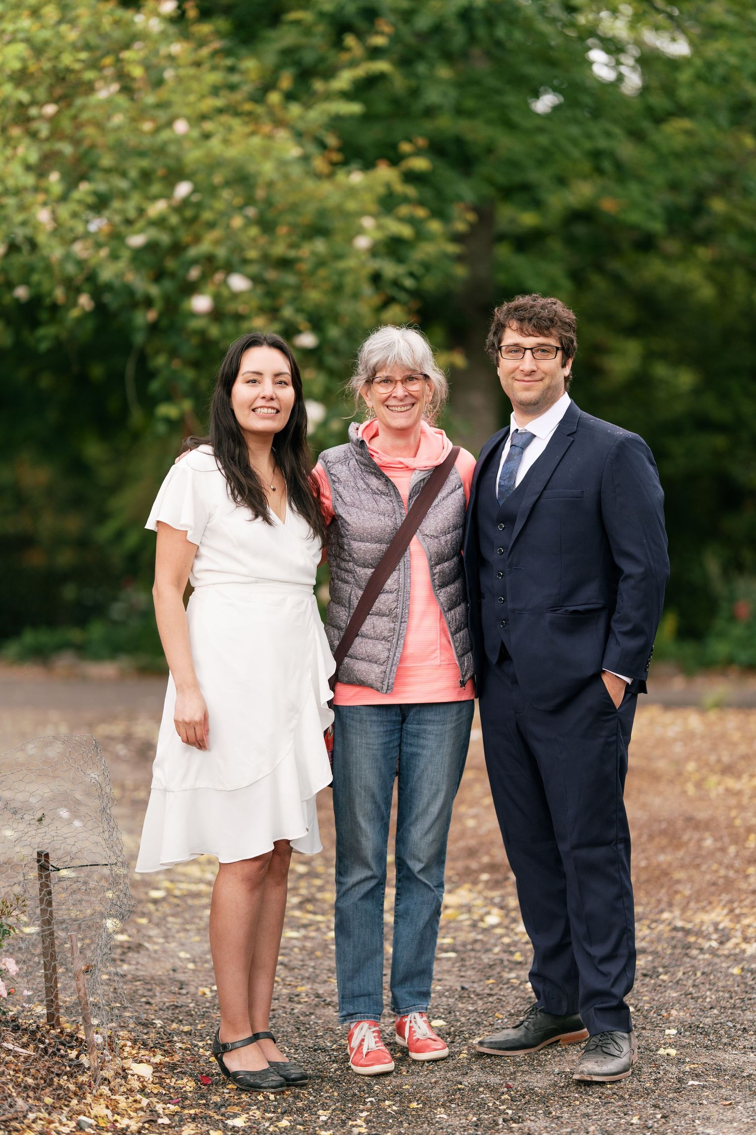 Bride and groom posing with wedding guest on a tree-lined garden path.