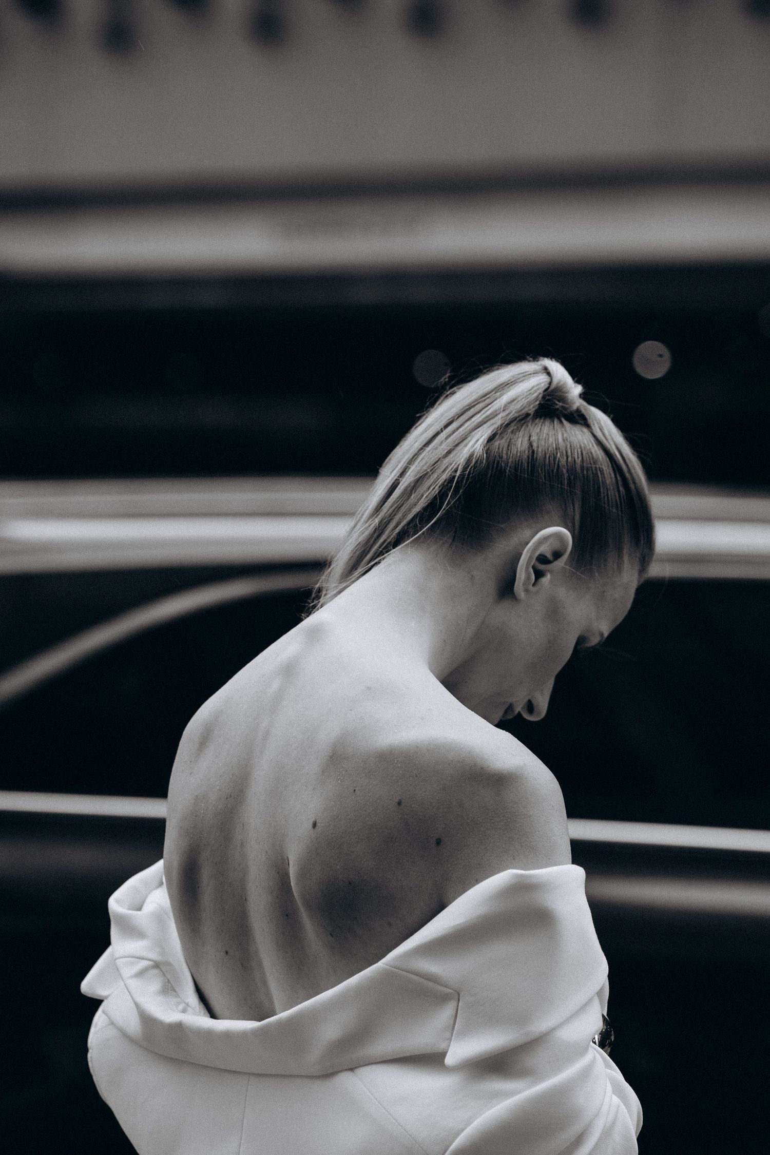 Black and white close-up of blonde woman with bare shoulder and white blazer pulled down in urban setting.