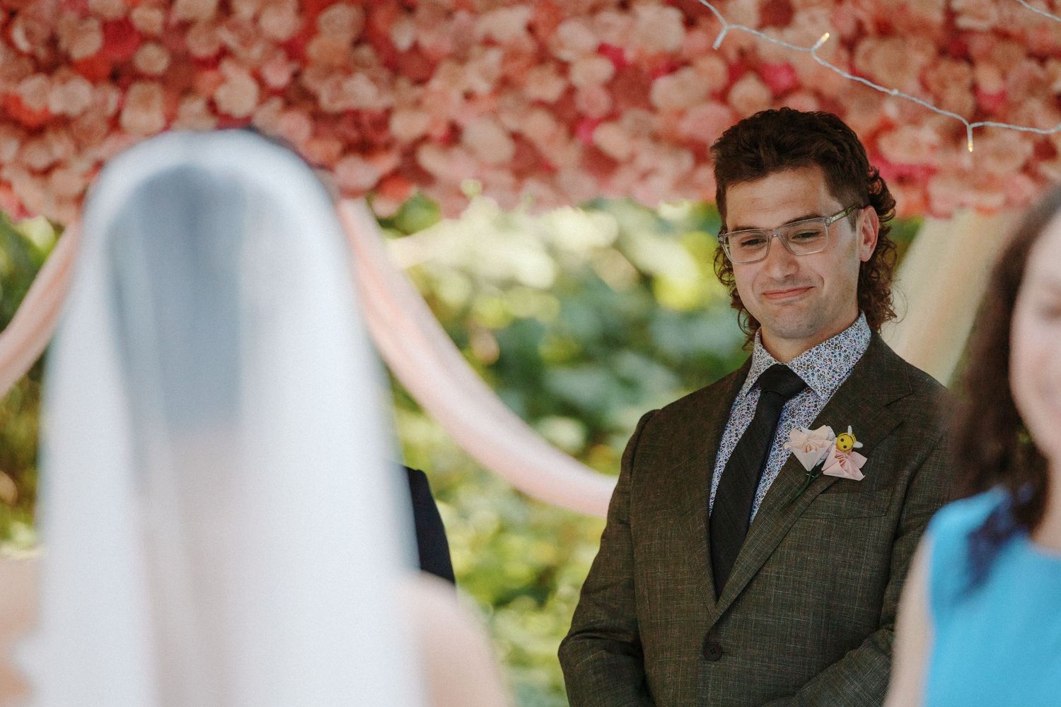 Emotional groom in grey suit and boutonniere gazes at bride during outdoor wedding ceremony.
