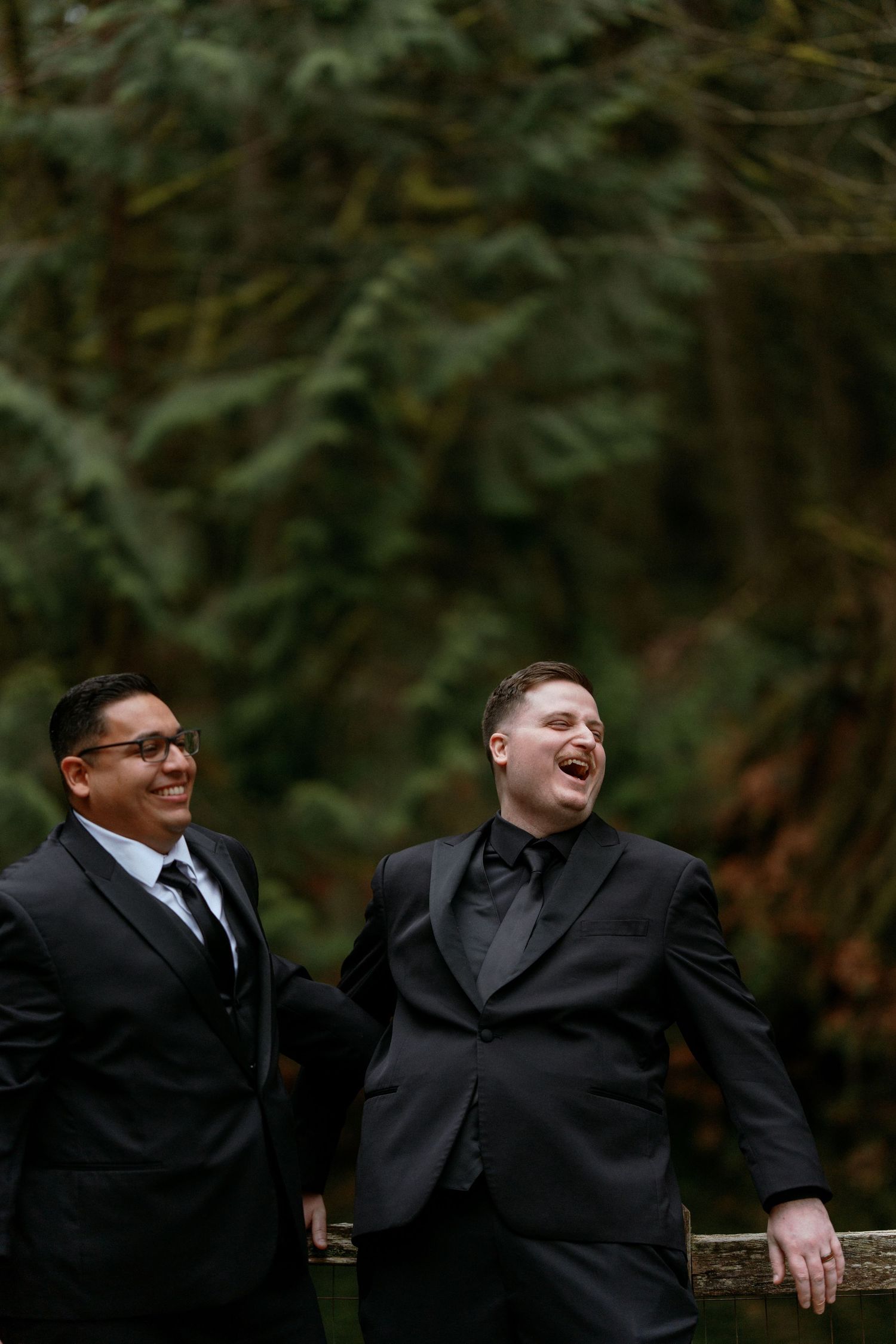 Two grooms in black tuxedos laughing joyfully outdoors in a lush forest wedding setting.