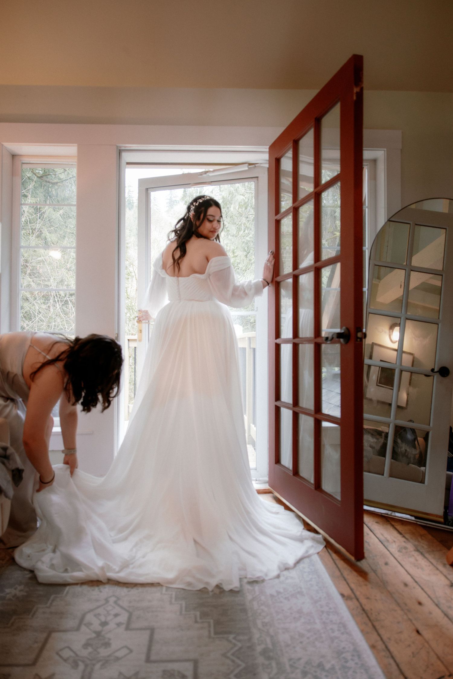 Bridesmaid adjusting bride's flowing white gown train near red French doors before wedding ceremony.