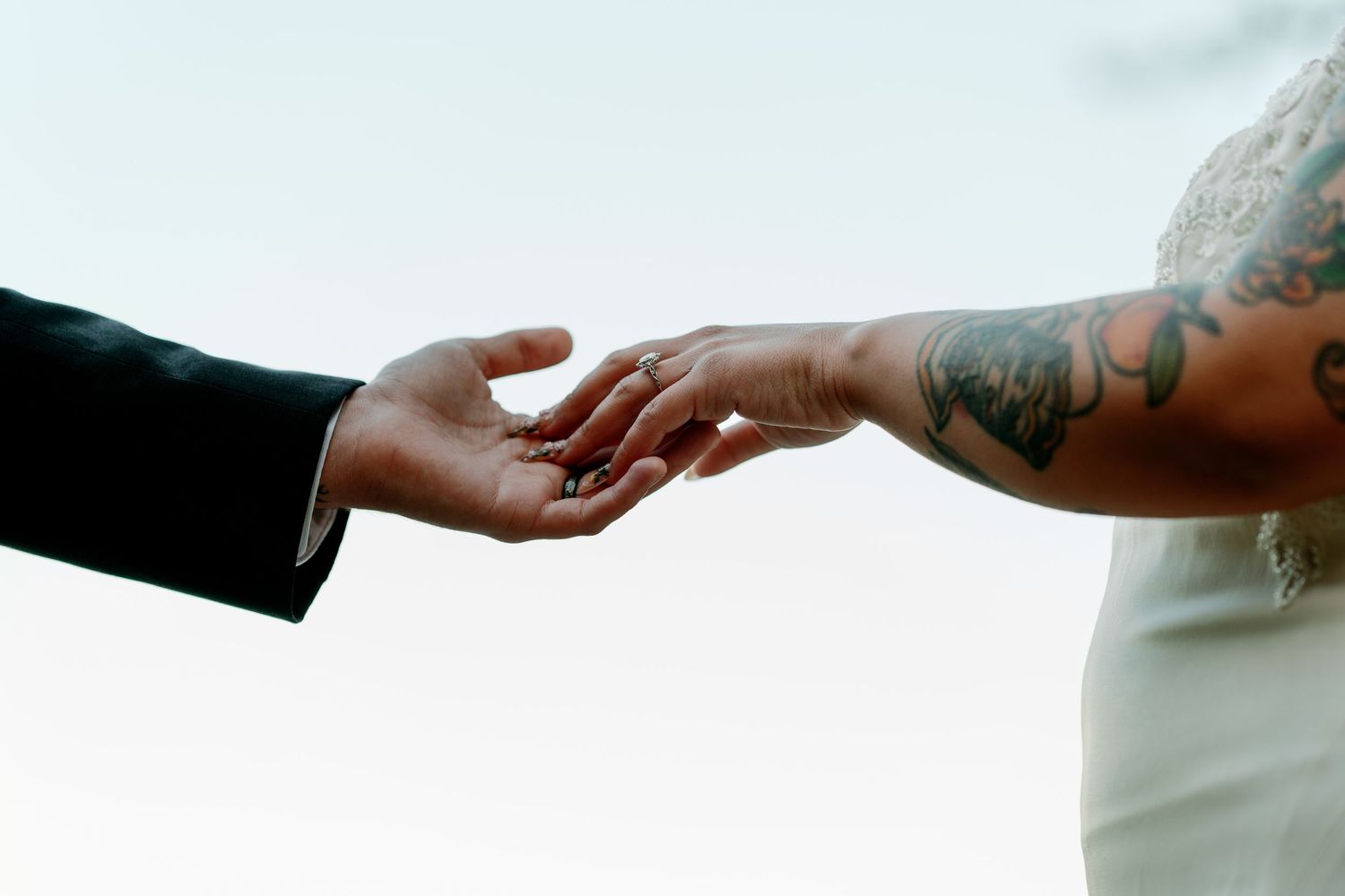 Close-up of bride and groom gently touching hands, showcasing wedding ring and bride's floral arm tattoo.