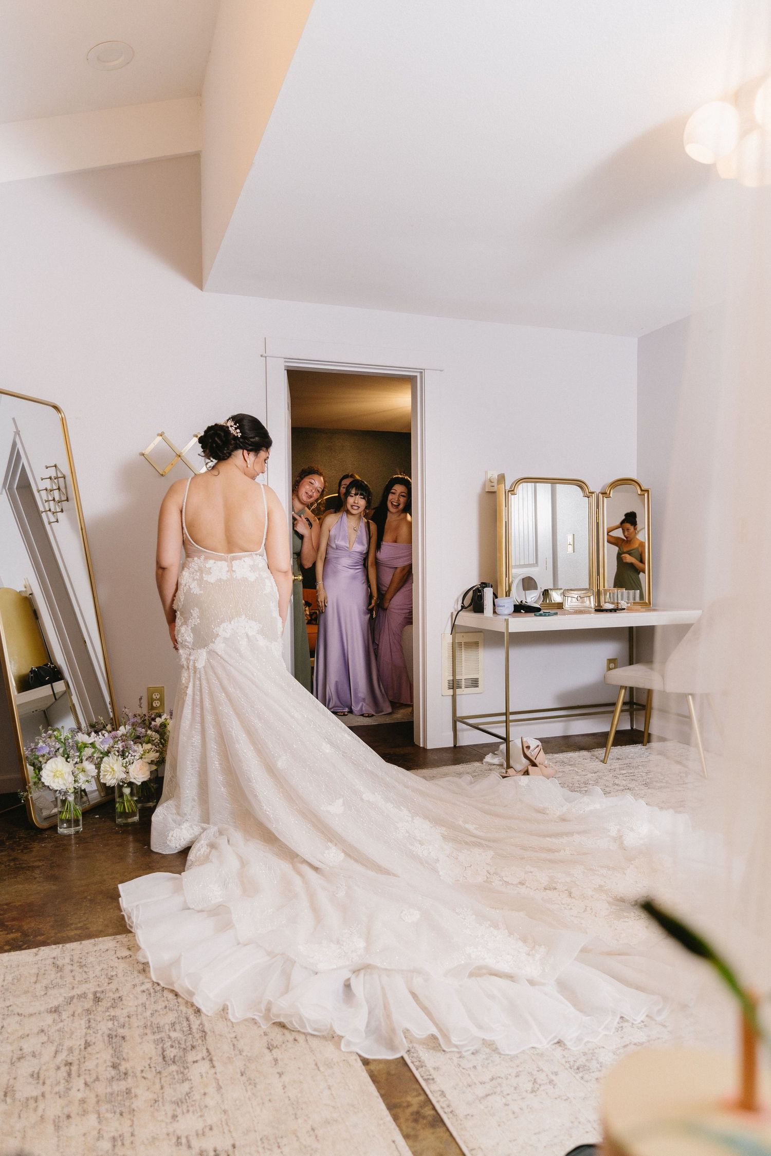 Bride in lace gown with long train as bridesmaids in lavender dresses look on.