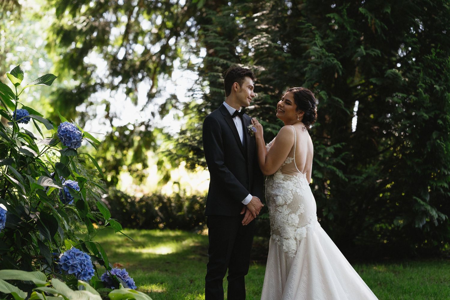 Bride and groom share a tender moment beside blooming blue hydrangeas in a sunlit outdoor garden setting.