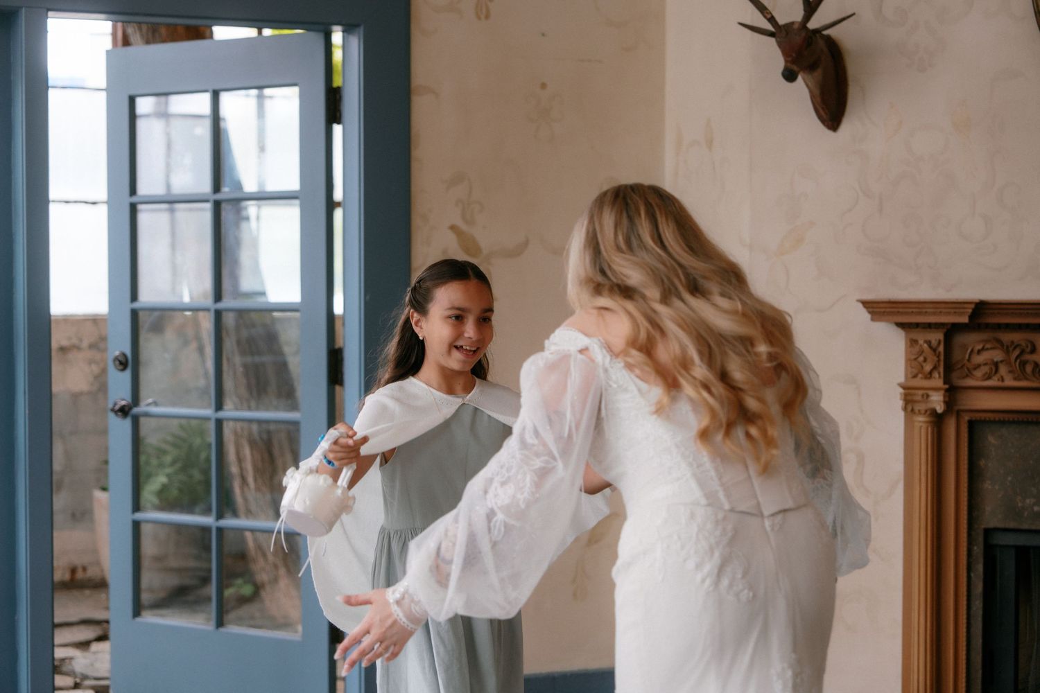 Bride in white lace dress sharing a joyful moment with a bridesmaid holding a garment bag indoors.