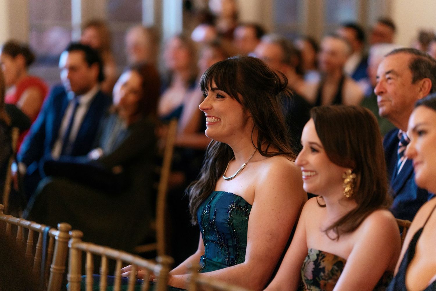 Wedding guests smile and laugh while seated at elegant indoor ceremony with chandeliers.