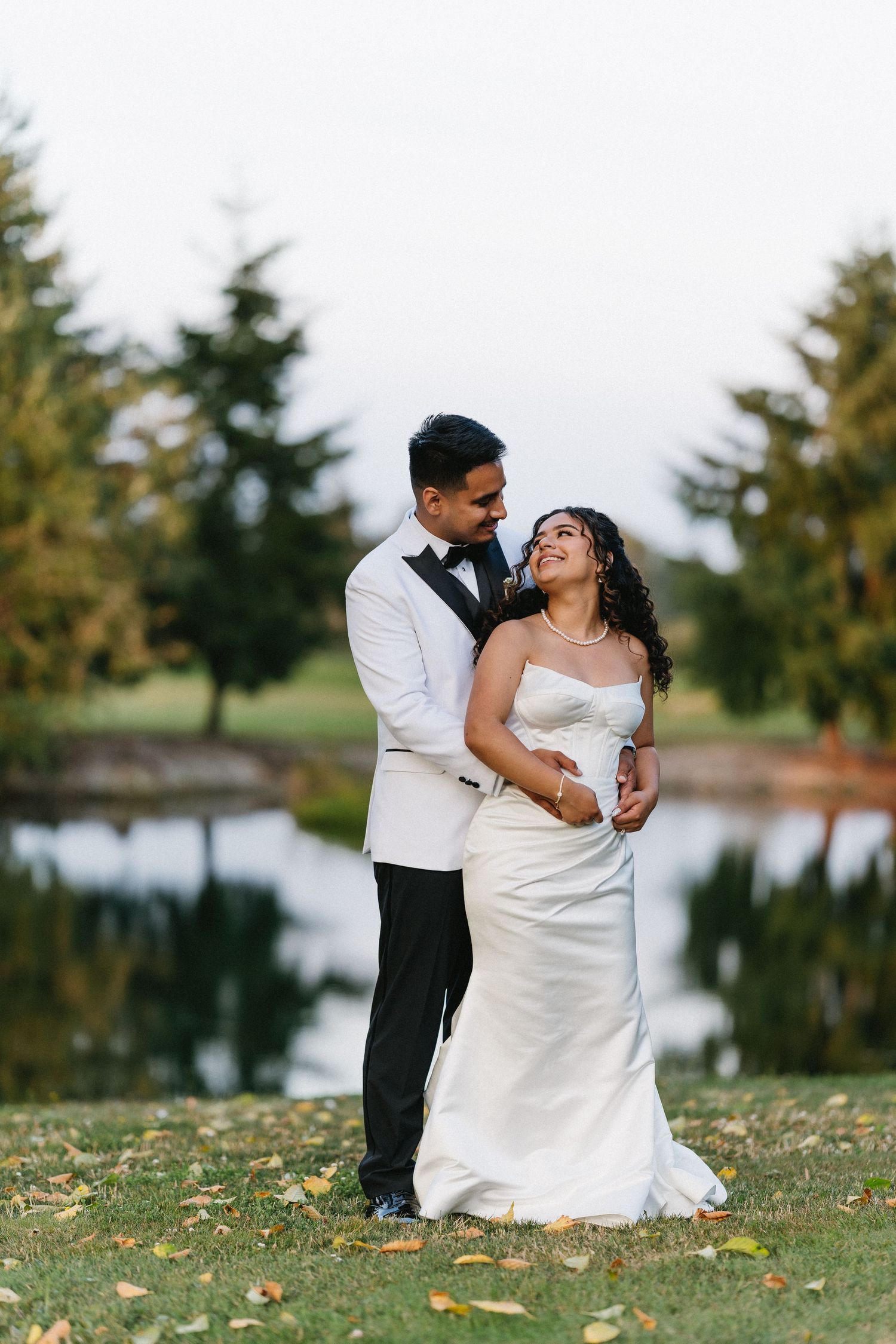 Bride and groom embrace near a reflective pond, she in a white gown, he in a white tuxedo jacket.