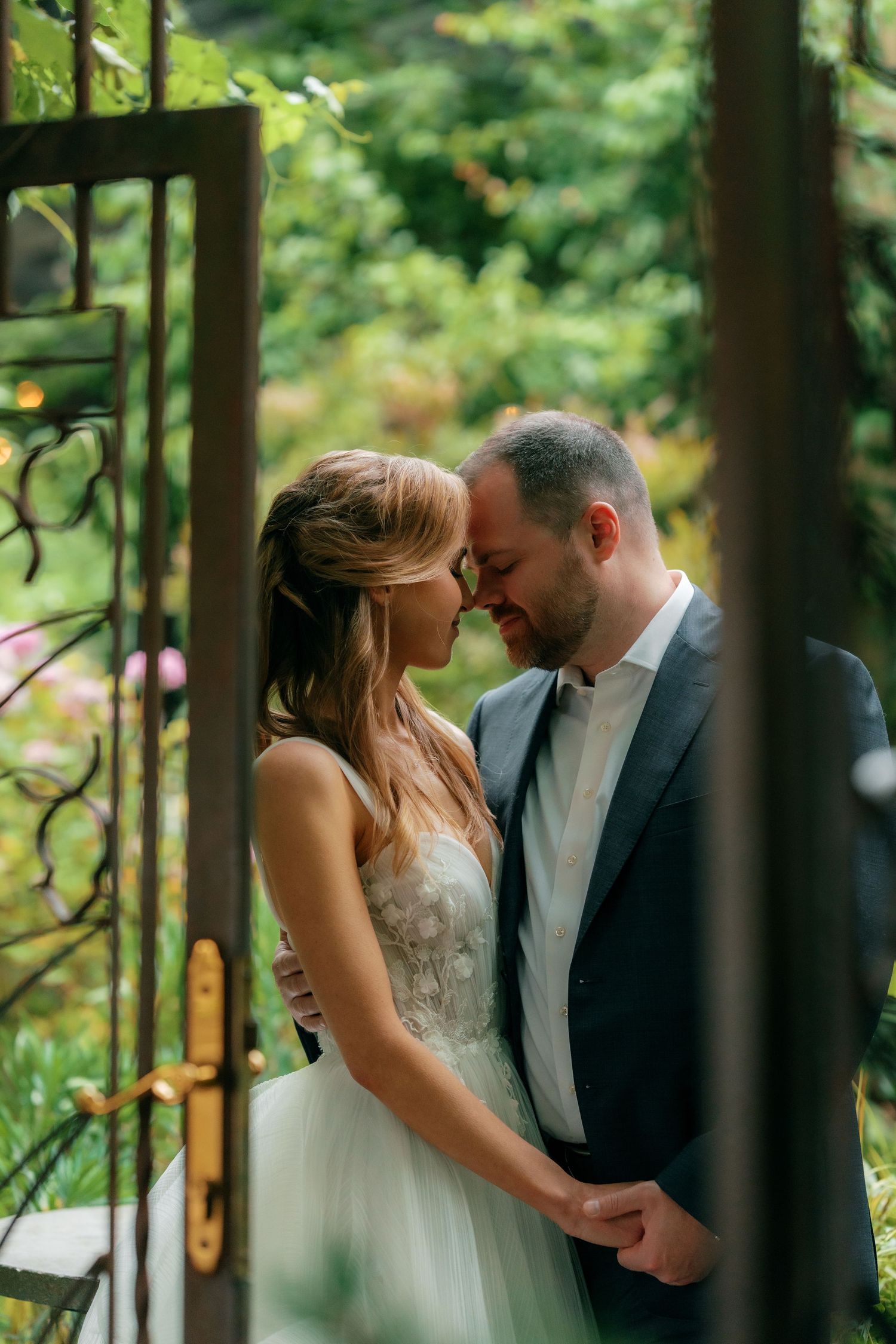 Bride and groom share an intimate moment framed by ornate iron doors in a garden.