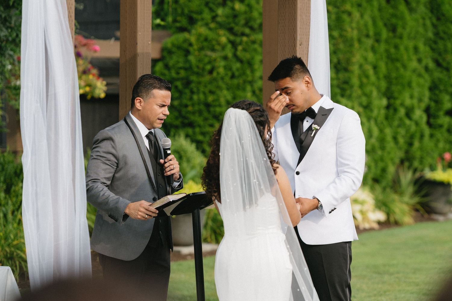 Emotional outdoor wedding ceremony with couple exchanging vows as officiant reads from book, bride in veil, groom in white tuxedo.