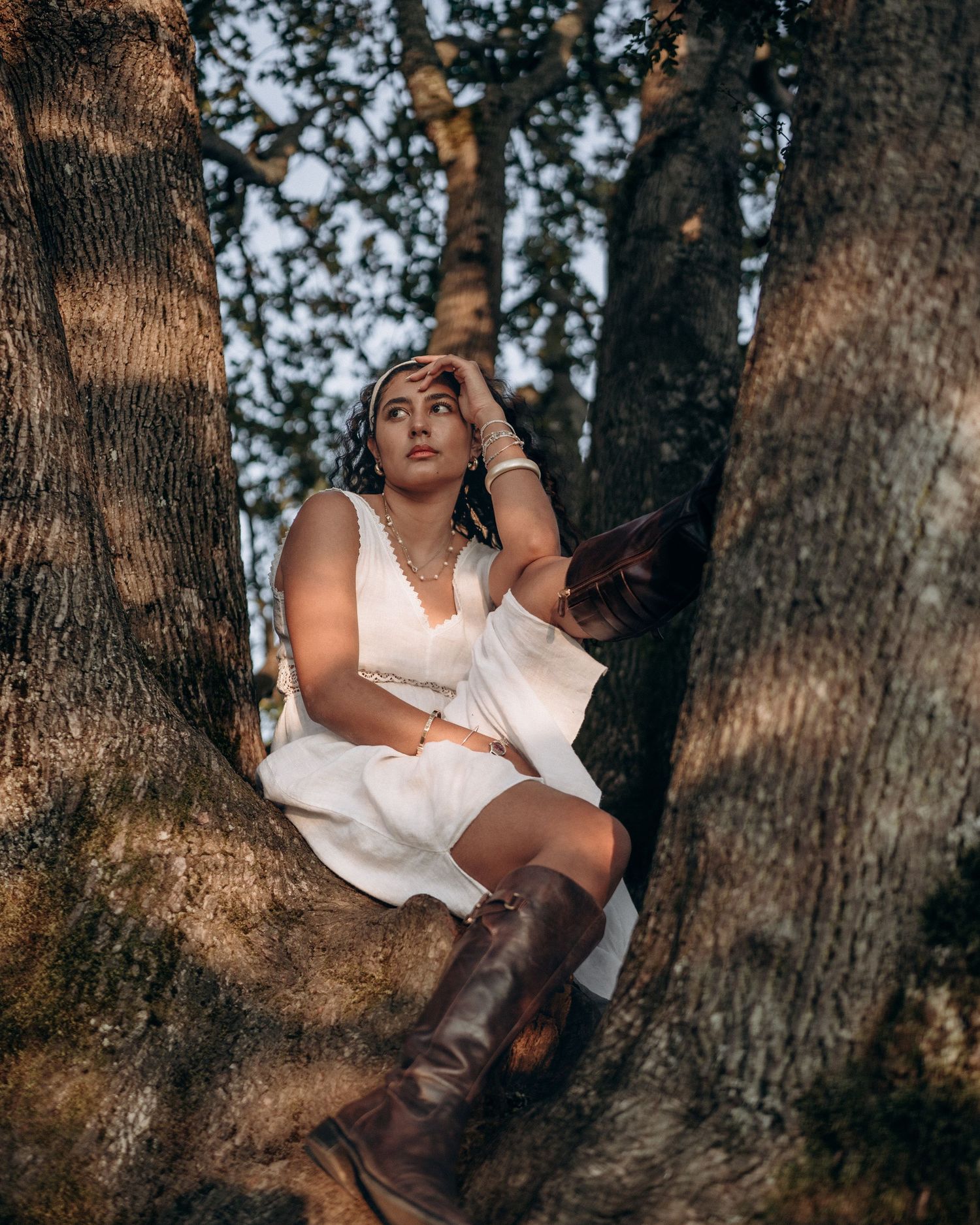 Woman in white dress and brown boots perched between tree trunks, looking upward in forest setting.