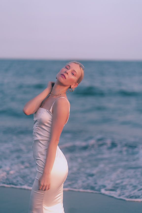 Elegant blonde woman in white satin slip dress poses by ocean waves at dusk.