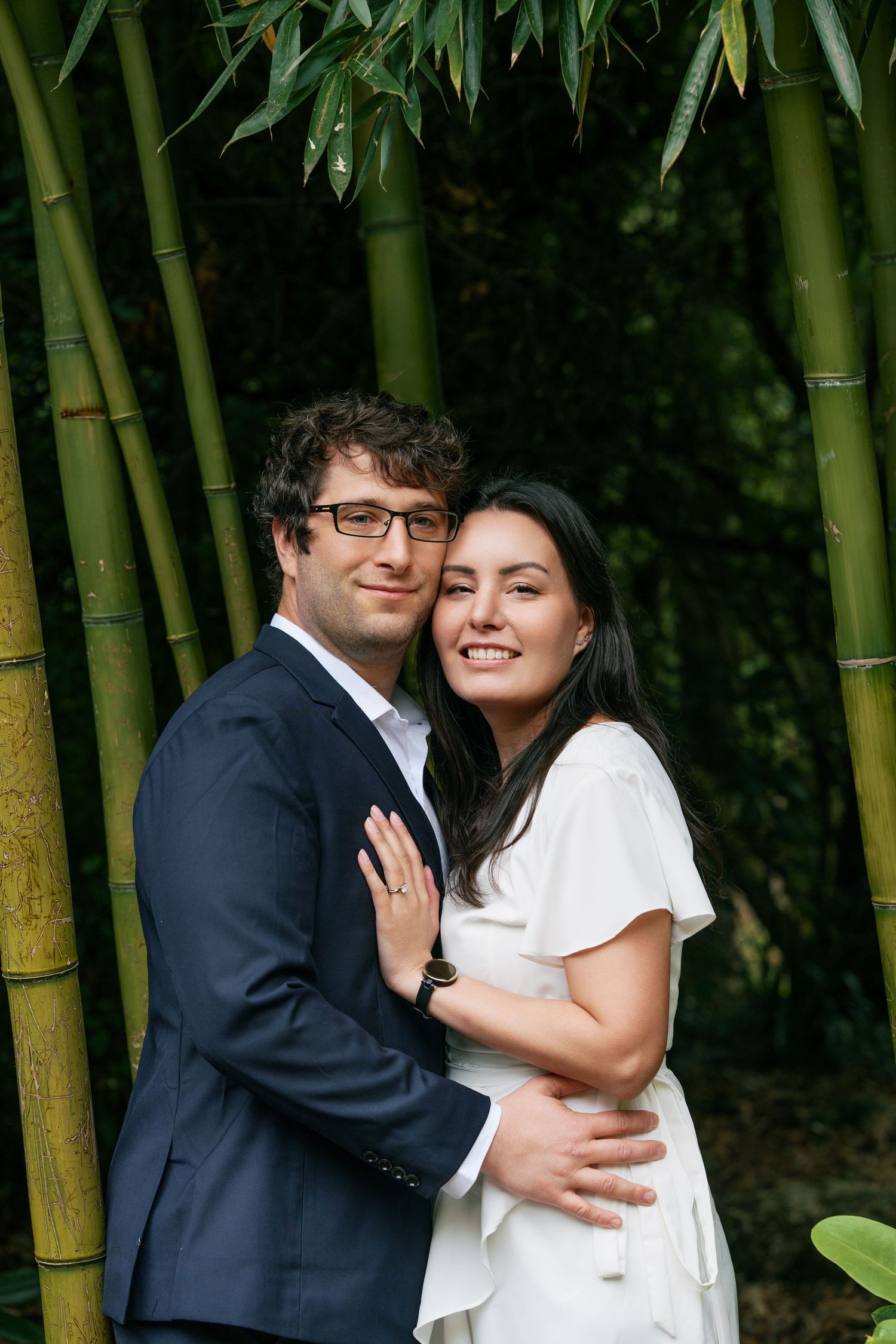Smiling couple posing together in front of tall bamboo stalks, man in navy suit and woman in white dress.
