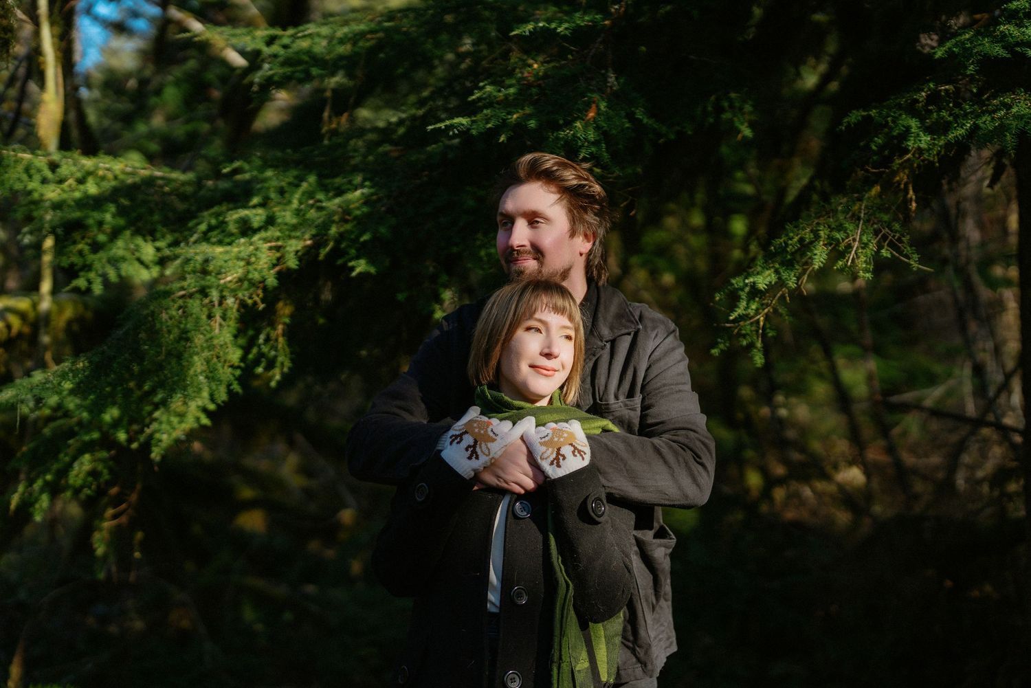 Man embracing woman from behind among green evergreen trees in a peaceful forest setting.