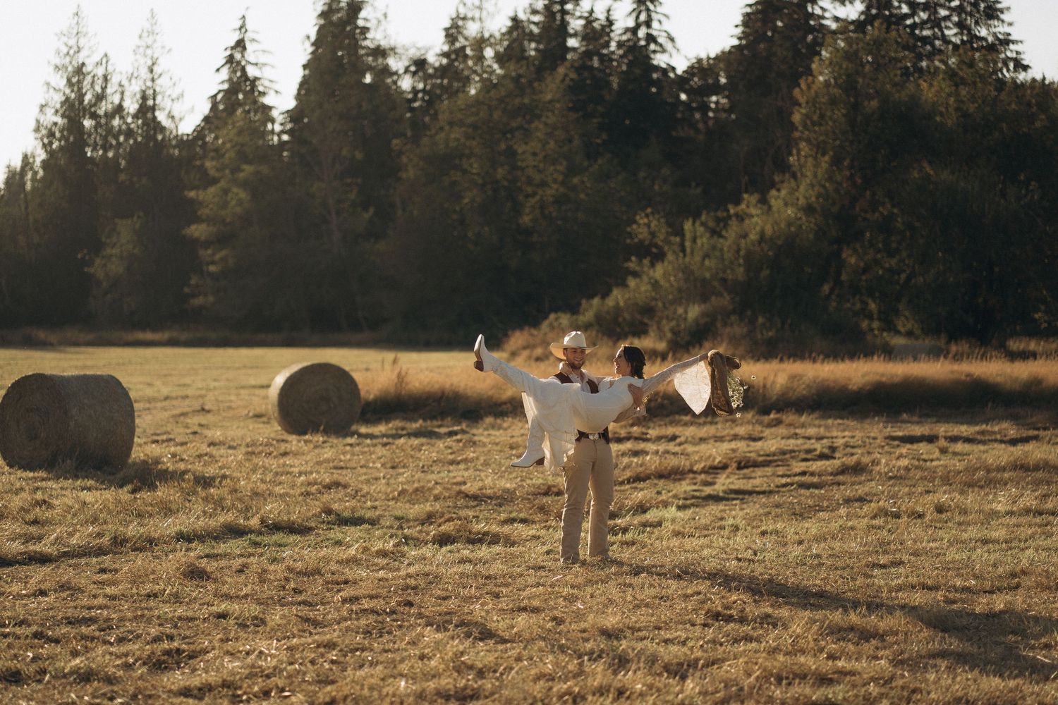 Couple spinning joyfully in golden field with hay bales and evergreen trees at sunset.