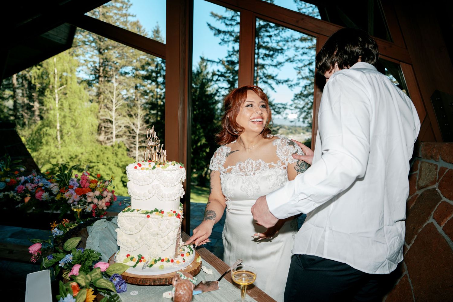 Bride and groom laughing joyfully while cutting wedding cake at sunny reception.