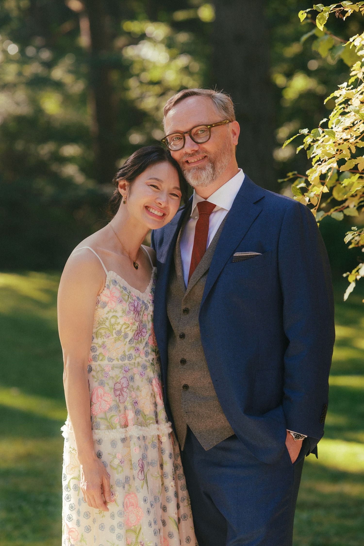 Smiling couple poses outdoors in wedding attire, woman in floral dress and man in navy suit with tie.