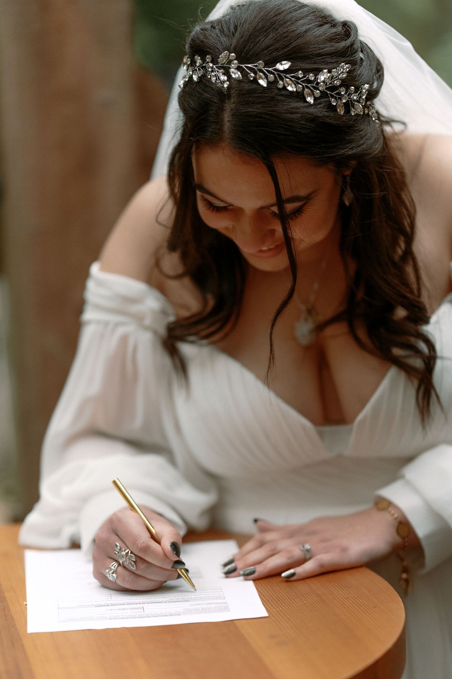 Bride in white off-shoulder gown signing wedding documents with gold pen, wearing floral crown and veil.