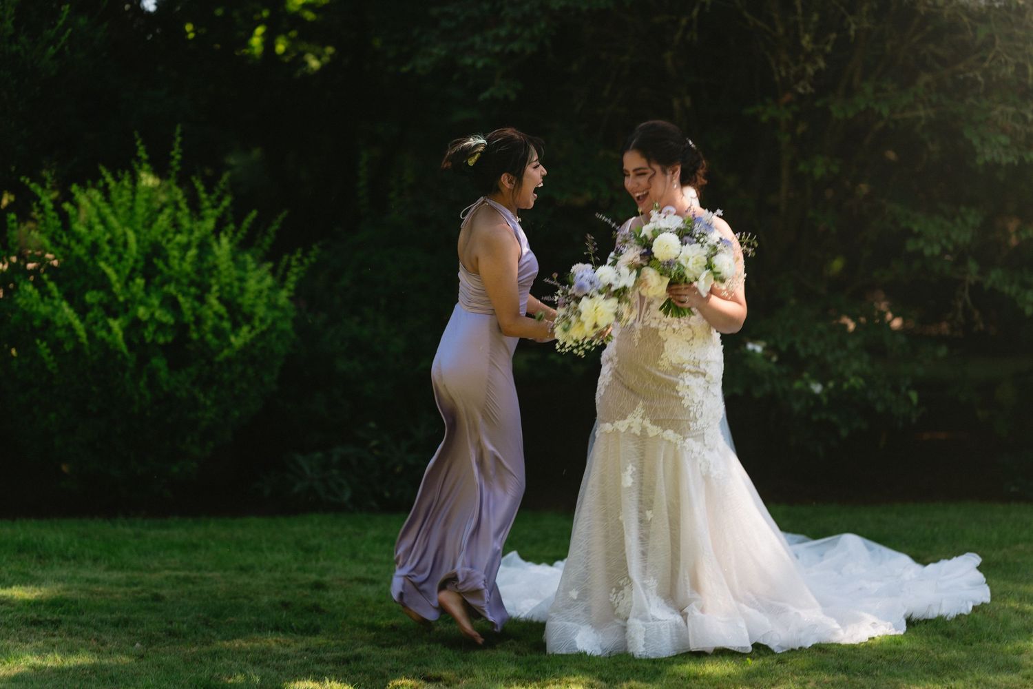 Bride in fitted lace wedding gown and bridesmaid in lavender dress sharing a moment in garden.