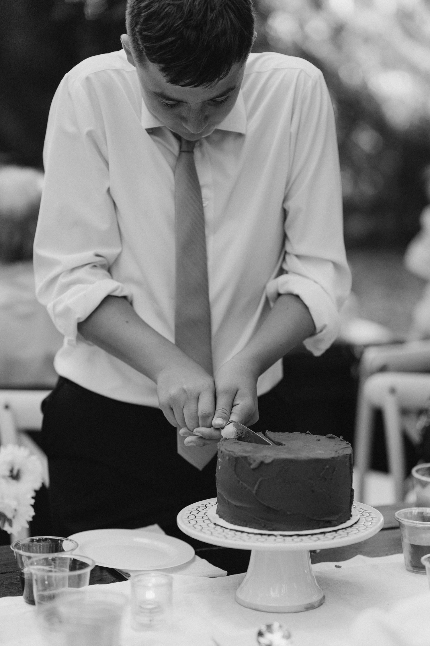 Black and white photo of young boy in tie carefully decorating a chocolate wedding cake.