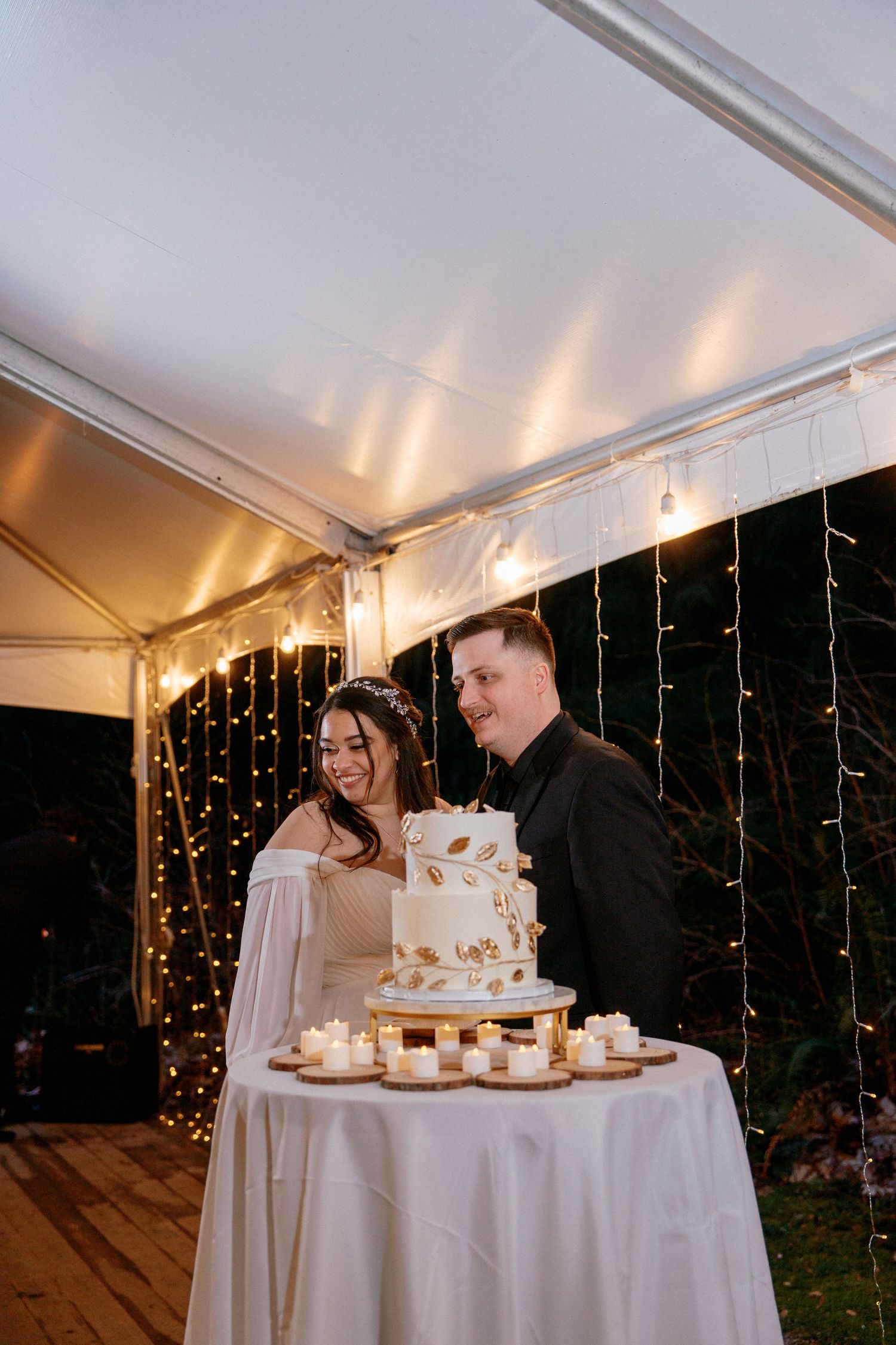 Bride and groom smile together beside a two-tier white and gold wedding cake decorated with candles under a tent.