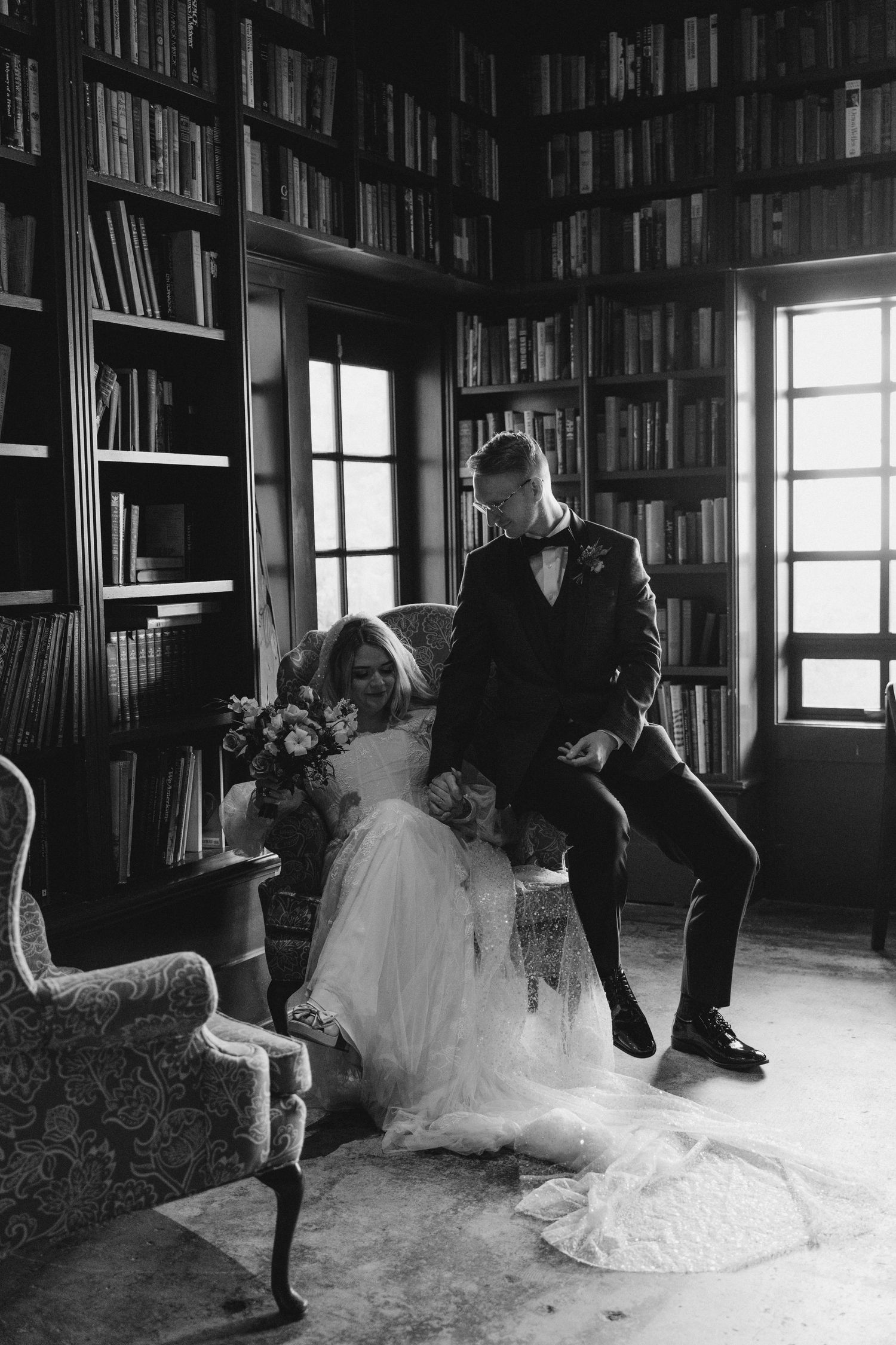 Bride and groom posing in a dramatic library with floor-to-ceiling bookshelves, black and white photo.