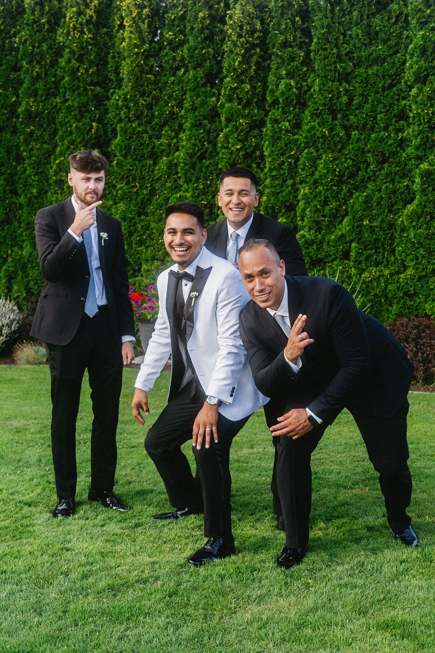 Four groomsmen in suits posing playfully on green lawn with tall hedges at wedding.